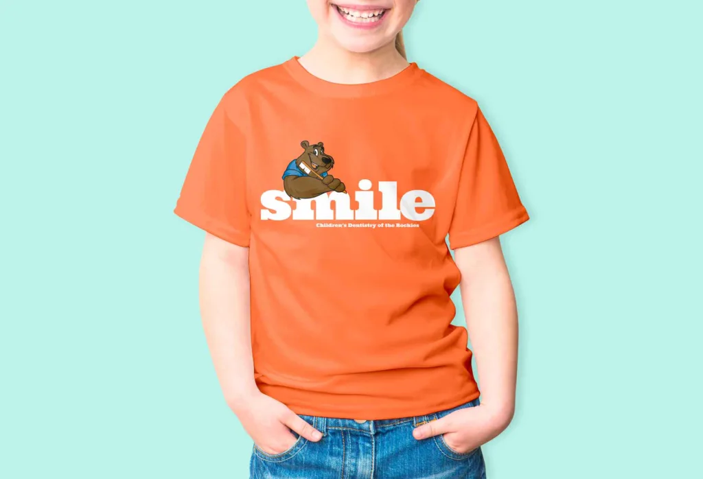 Girl smiling with hands in pockets wearing pediatric dentist t-shirt of a bear with the word smile in large print